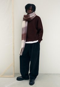 Mörkröd stickad tröja, oversize svarta kordbyxor, och en mönstrad scarf i ockra, krämfärg och mauve. Klumpiga svarta skor med metalldetaljer.