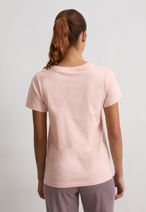 Vrouw met lang bruin haar in een paardenstaart, gekleed in een eenvoudig lichtroze T-shirt met korte mouwen en gemusterde broek, met haar rug naar de camera tegen een effen achtergrond.