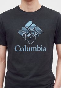 Άτομο που φοράει μαύρο μπλουζάκι Columbia με μεγάλο ανοιχτό μπλε λογότυπο και κείμενο στο στήθος.