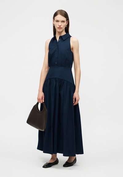 Γυναίκα με αμάνικο φόρεμα σε σκούρο μπλε με κουμπιά, κρατώντας σκούρα καφέ τσάντα χειρός, στέκεται με μαύρα φλατ παπούτσια σε λευκό φόντο.