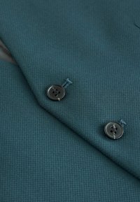 Dunkeltürkiser, strukturierten Stoff mit dezentem Muster, versehen mit zwei schwarzen Knöpfen und kontrastierenden Nähakzenten am Kragen und Revers.