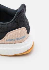 Gros plan sur le talon d'une basket noire et beige avec le texte bleu clair « ultra boost », une semelle intermédiaire blanche texturée et une semelle extérieure en caoutchouc naturel sur fond blanc.