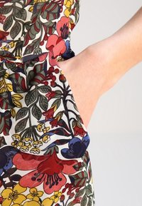 Tkanina w kwiaty ma wielokolorowe kwiaty w odcieniach czerwieni, niebieskiego, żółtego i zielonego. Ręka sięga do bocznej kieszeni.