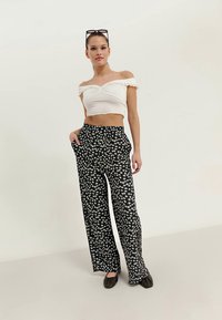 Kvinne iført off-shoulder hvit crop top, svarte vide bukser med hvitt blomstermønster, svarte flate sko og solbriller på hodet.