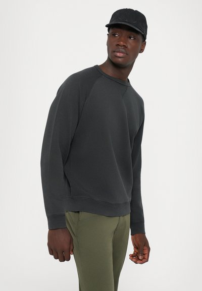 Mann som har på seg svart caps, mørk grå sweatshirt, og olivengrønne bukser, og ser over venstre skulder mot en ensfarget lys bakgrunn.