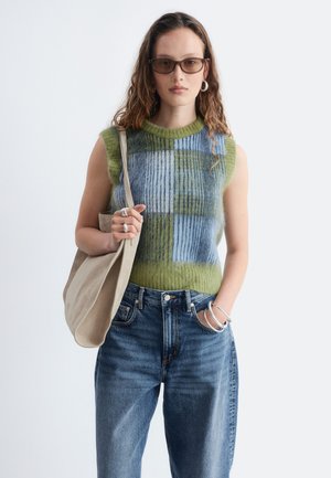 Jeune femme aux longs cheveux bouclés portant des lunettes de soleil, un haut sans manches en maille à motif vert et bleu, un jean bleu, des bijoux en argent et un sac beige.