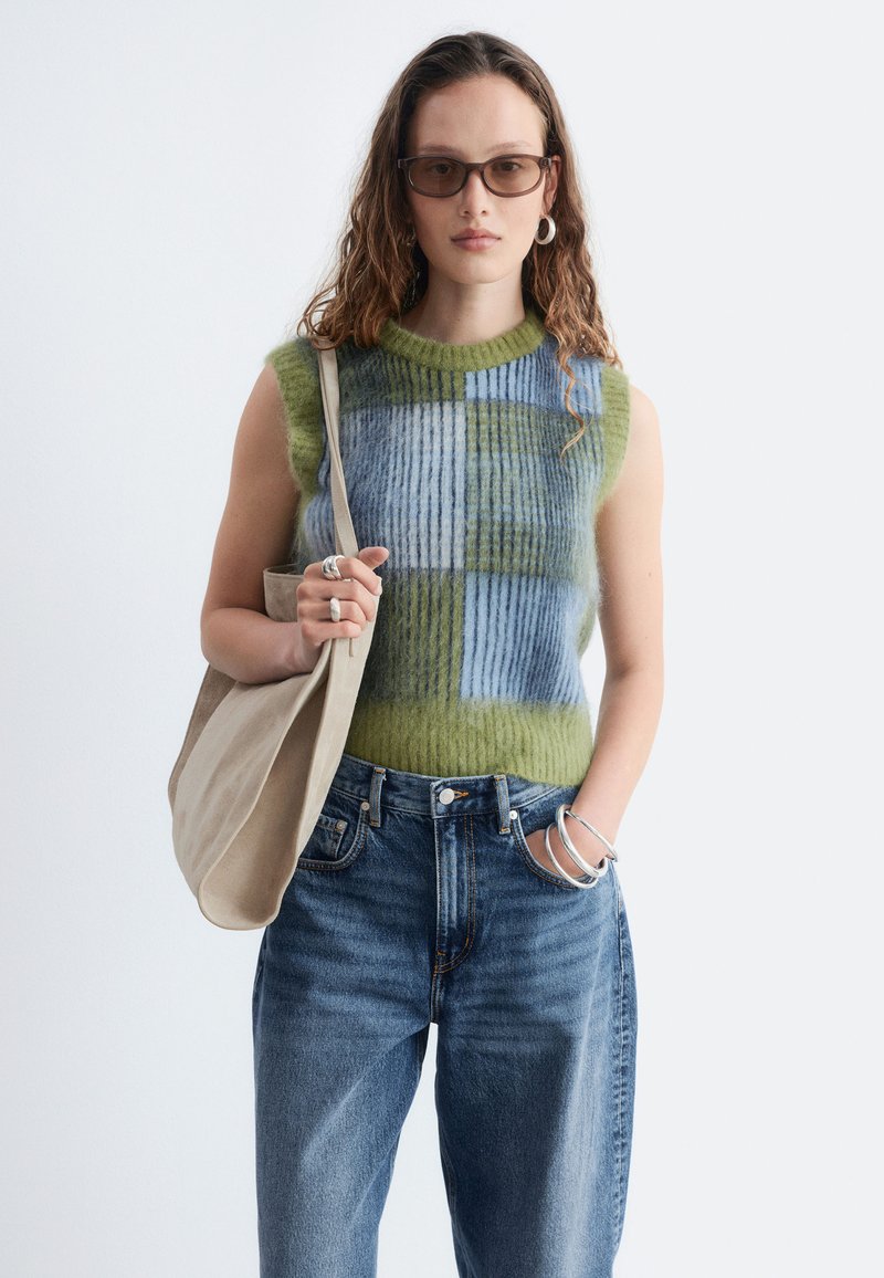 Jeune femme aux longs cheveux bouclés portant des lunettes de soleil, un haut sans manches en maille à motif vert et bleu, un jean bleu, des bijoux en argent et un sac beige.