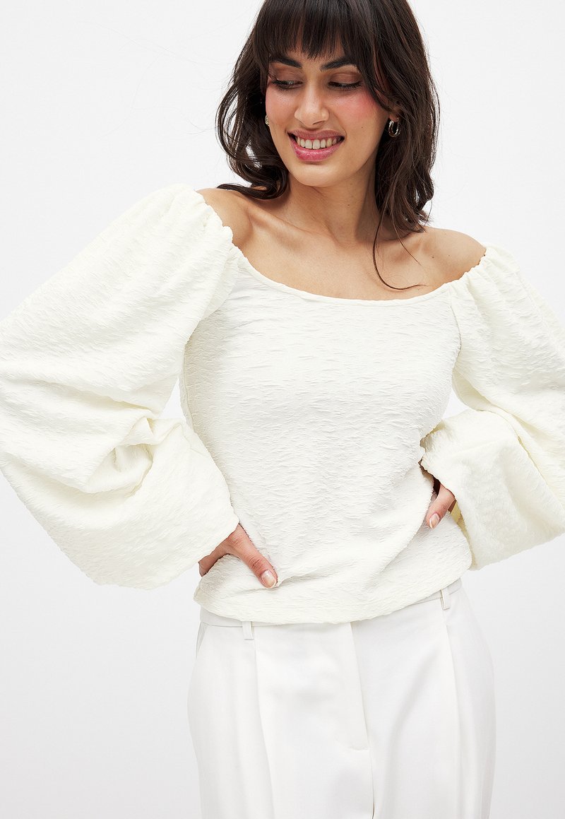 Witte blouse met een off-shoulder halslijn, textuurstof en pofmouwen. Gecombineerd met lichtgekleurde broeken.