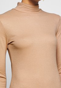 Ribattu turtleneck yläosa pehmeässä beigen värisessä kankaassa, jossa on tiukka muotoilu, pitkät hihat ja korkea kaulus. Tasainen rakenne hienovaraisella kiillolla.