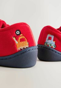 Chaussons en polaire rouge avec semelles en caoutchouc bleu marine. Chacun a des motifs de véhicules brodés : un camion jaune et un train multicolore.