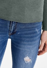 Grünes, geripptes Langarmoberteil, kombiniert mit ausgefransten blauen Jeans aus Denim, die ein sichtbares Loch am linken Oberschenkel und dunkle Nähte aufweisen.