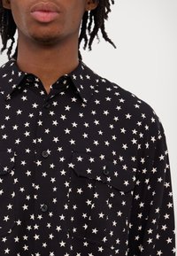 Černá košile s bílým hvězdným vzorem, zapínání na knoflíky, dvě náprsní kapsy s klopami, krátké rukávy a strukturovaná tkanina.
