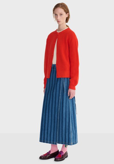Femme portant un cardigan rouge boutonné, un haut blanc, une longue jupe en denim rayée bleue, des chaussettes roses avec un croissant vert, et des mocassins foncés à pompons.