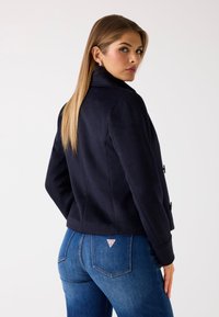 Chaqueta azul marino de corte doble con solapa, hecha de material suave. Presenta detalles de botones y un ajuste entallado, llevada sobre jeans azules.