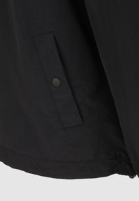 Czarna lekka kurtka o gładkiej fakturze, wyposażona w boczną kieszeń z okrągłym zamknięciem na guzik. Prosty design, bez wzorów i ozdób.