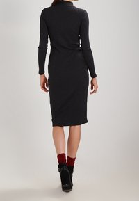 Svart ribbad midiklänning med långa ärmar, åtsittande form och hög krage. Modellen bär röda strumpor och kraftiga svarta klackskor.