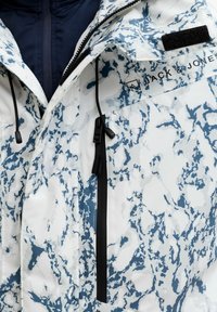 Veste blanche avec un motif abstrait bleu, zips noirs et cordon de serrage. Elle présente un col montant et un logo sur le côté supérieur gauche.