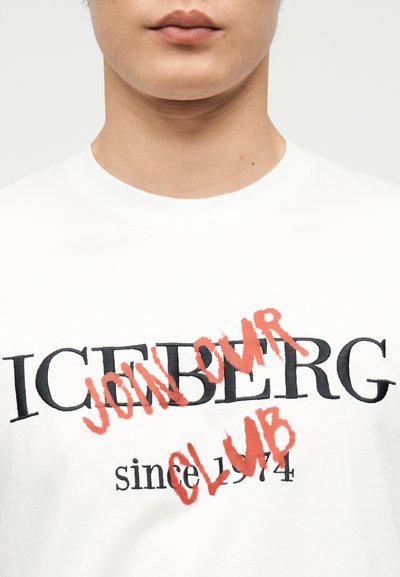 Κοντινό πλάνο ενός ατόμου που φοράει λευκό μπλουζάκι με μαύρο κείμενο «ICEBERG» και κόκκινη μπογιά ψεκασμένη με το κείμενο «JON OUR CLUB» πάνω του, με μαύρο κείμενο «since 1974» από κάτω.