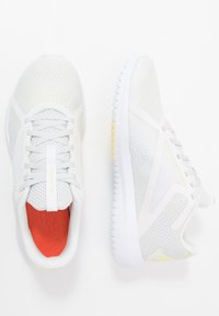 Bílé sportovní tenisky s prodyšným síťovinovým svrškem, texturovanými vzory, bílou gumovou podrážkou a oranžovým detailem na stélce.