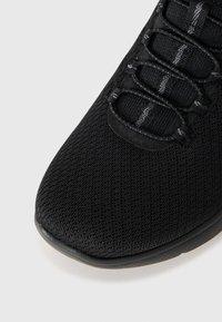 Chaussure de sport noire avec une tige en mesh texturé, dotée de lacets élastiques et d'un col rembourré. Bout arrondi et semelle en caoutchouc durable.