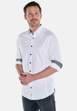 Man with short brown hair wearing white button-up shirt with black buttons and patterned rolled-up sleeves, standing with hands clasped.