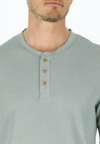 Vaaleanvihreä henley-paita, joka on valmistettu teksturoidusta kankaasta ja jossa on kolme ruskeaa nappia ja pyöreä pääntie.