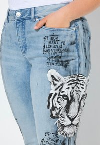 Jeans de denim azul claro con un estampado de tigre en el muslo y gráficos de texto. Diseñados con una silueta ajustada y bolsillos.