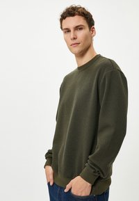Khaki groene geribde sweatshirt met een ronde hals en lange mouwen, voorzien van elastische manchetten en een relaxed fit, gecombineerd met blauwe spijkerbroek.