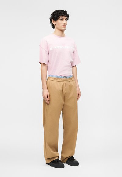 Άτομο όρθιο, φοράει ροζ ανοιχτό oversized μπλουζάκι, μπεζ φαρδιά παντελόνια και μαύρα αθλητικά παπούτσια μπροστά σε απλό φόντο.