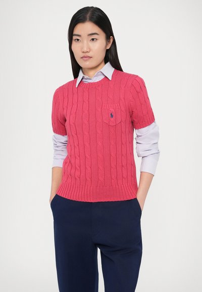 Ροζ πλεκτή μπλούζα με σύντομο μανίκι, που διαθέτει τσέπη και μικρό κεντημένο λογότυπο. Φοριέται πάνω από ένα γαλάζιο πουκάμισο με γιακά.