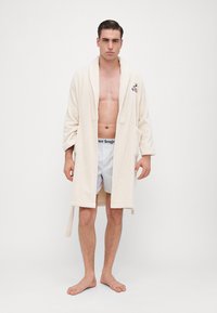 Κρεμ χρώματος μπουρνούζι με υφή, μαλακό ύφασμα, που διαθέτει γιακά σαλονιού και πλάγιες τσέπες, φορεμένο πάνω από ανοιχτόχρωμα σορτσάκια.