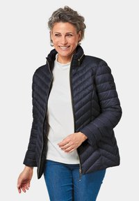 Doudoune matelassée bleu marine avec un col montant, fermeture éclair et poches latérales ; présente un motif de couture linéaire et des accents dorés ; matière légère.