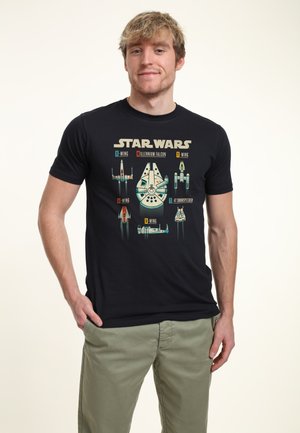 Ung mand iført en sort Star Wars T-shirt med mærkede illustrationer af Millennium Falcon, X-Wing, Y-Wing, A-Wing, B-Wing og 47 Snowspeeder.