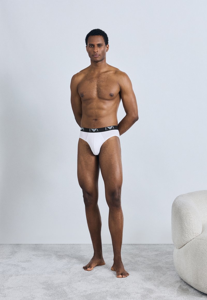 Slip bianco da uomo con elastico in vita nero decorato con dettagli logo. Modello in piedi a piedi nudi su un tappeto grigio chiaro.