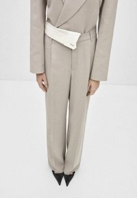 Costum bej, croit cu un sacou ajustat și pantaloni largi. Un brâu alb în contrast adaugă detalii. Pantofi negri cu vârful ascuțit completează lookul.