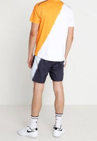 Homme portant une chemise à manches courtes orange et blanche, un short noir et blanc, des baskets blanches Nike et des chaussettes blanches, debout devant un fond uni.