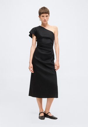 Henkilö seisoo päällään musta olkapäältä paljastava midihame, jossa on kerätty hiha, ja mustat Mary Janen tasapohjakengät valkoisella taustalla.