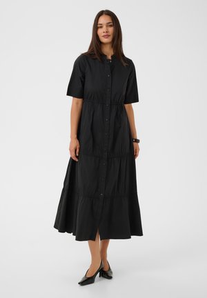 Femme debout portant une robe midi noire boutonnée avec manches courtes et des talons bas noirs sur un fond clair uni.