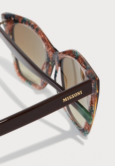 Hexagonale Sonnenbrille mit einem multikolorierten Glitzerrahmen, braunen Bügeln und grün getönten Gläsern. Goldene "MISSFONI"-Markierung auf dem Bügel.