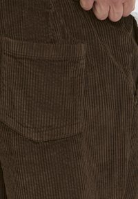 Pantaloni marroni in velluto a coste con texture a coste verticali e una tasca a toppa laterale, che mostrano un tessuto morbido e resistente.