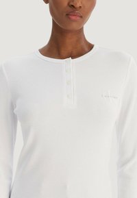 Bílý žebrovaný top s dlouhým rukávem a kulatým výstřihem, zdobený zapínáním na knoflíky a malým vyšívaným logem na levé straně.