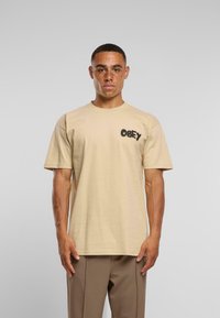 Muž s pihami, oblečený v béžovém tričku s logem "Obey" a hnědých kalhotách, stojící na jednobarevném světle šedém pozadí.