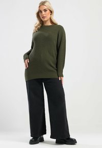 Olivgrüner Strickpullover mit weitem Ausschnitt und gerippter Textur, kombiniert mit hochgeschnittenen schwarzen weit geschnittenen Hosen und klobigen schwarzen Stiefeln.