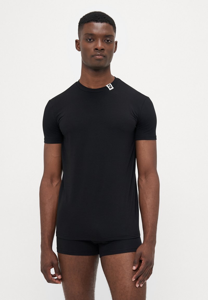 Mann in einem enganliegenden schwarzen T-Shirt ohne Aufdruck mit einem kleinen weißen Fleck am Kragen und passenden schwarzen Shorts, der vor einem weißen Hintergrund steht.