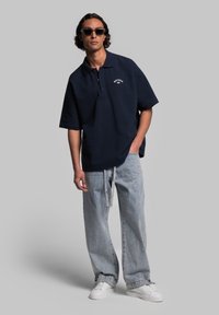 Granatowa koszulka polo z krótkim rękawem i kołnierzykiem, z małym białym logo. Noszona z jasnoniebieskimi, luźnymi dżinsami i białymi trampkami.