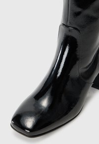 Botim de tornozelo preto em verniz com bico quadrado, design elegante e salto ligeiramente elevado. Textura suave com detalhes de costura mínimos.