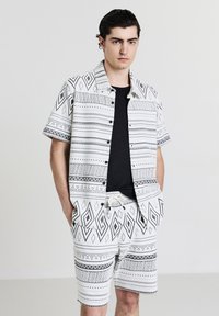 Jeune homme portant une chemise à manches courtes à motif géométrique noir et blanc assortie à un short, avec un t-shirt noir en dessous, les mains dans les poches.
