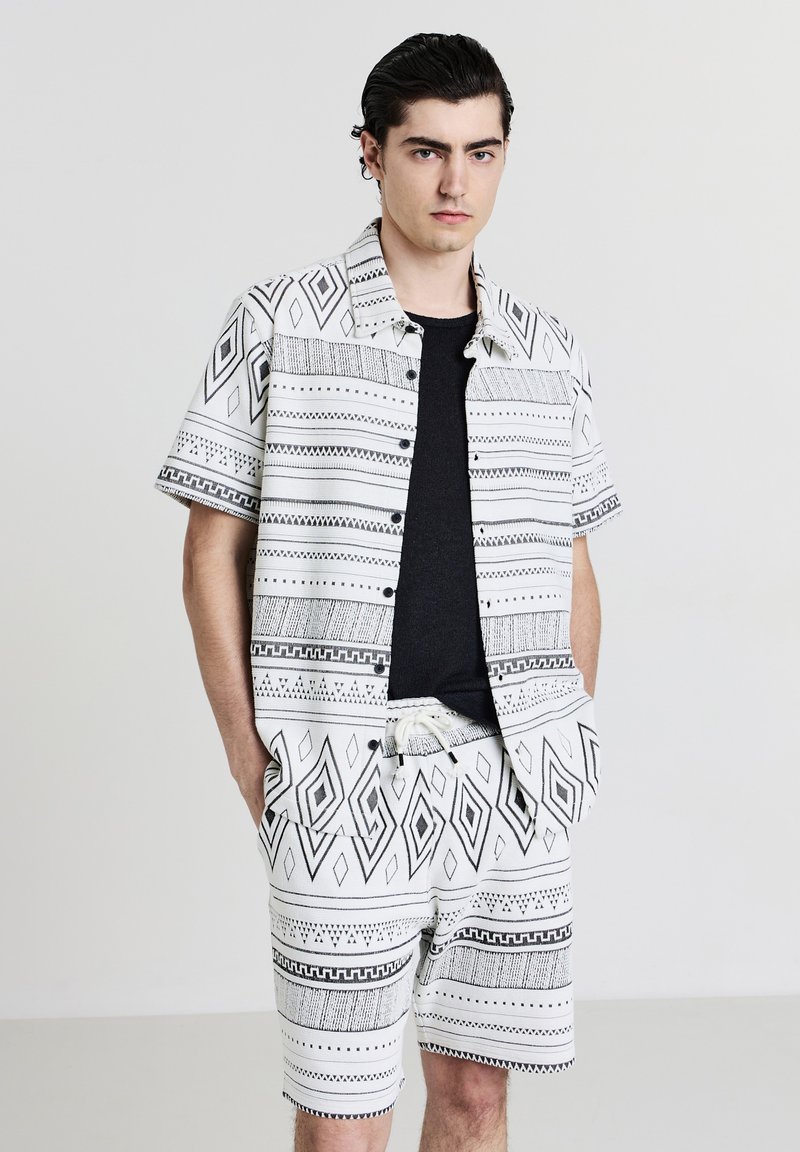 Jeune homme portant une chemise à manches courtes à motif géométrique noir et blanc assortie à un short, avec un t-shirt noir en dessous, les mains dans les poches.