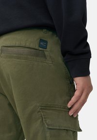 Olivgrüne Cargo-Hose mit einer Gesäßtasche, strukturiertem Stoff und einem kleinen Logo-Patch. Jede Seite hat Utility-Taschen für zusätzlichen Stauraum.