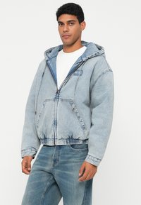 Hombre que lleva una sudadera de mezclilla azul claro con cremallera en el frente y bolsillo tipo canguro, combinada con jeans azules y una camiseta blanca, de pie contra un fondo blanco.